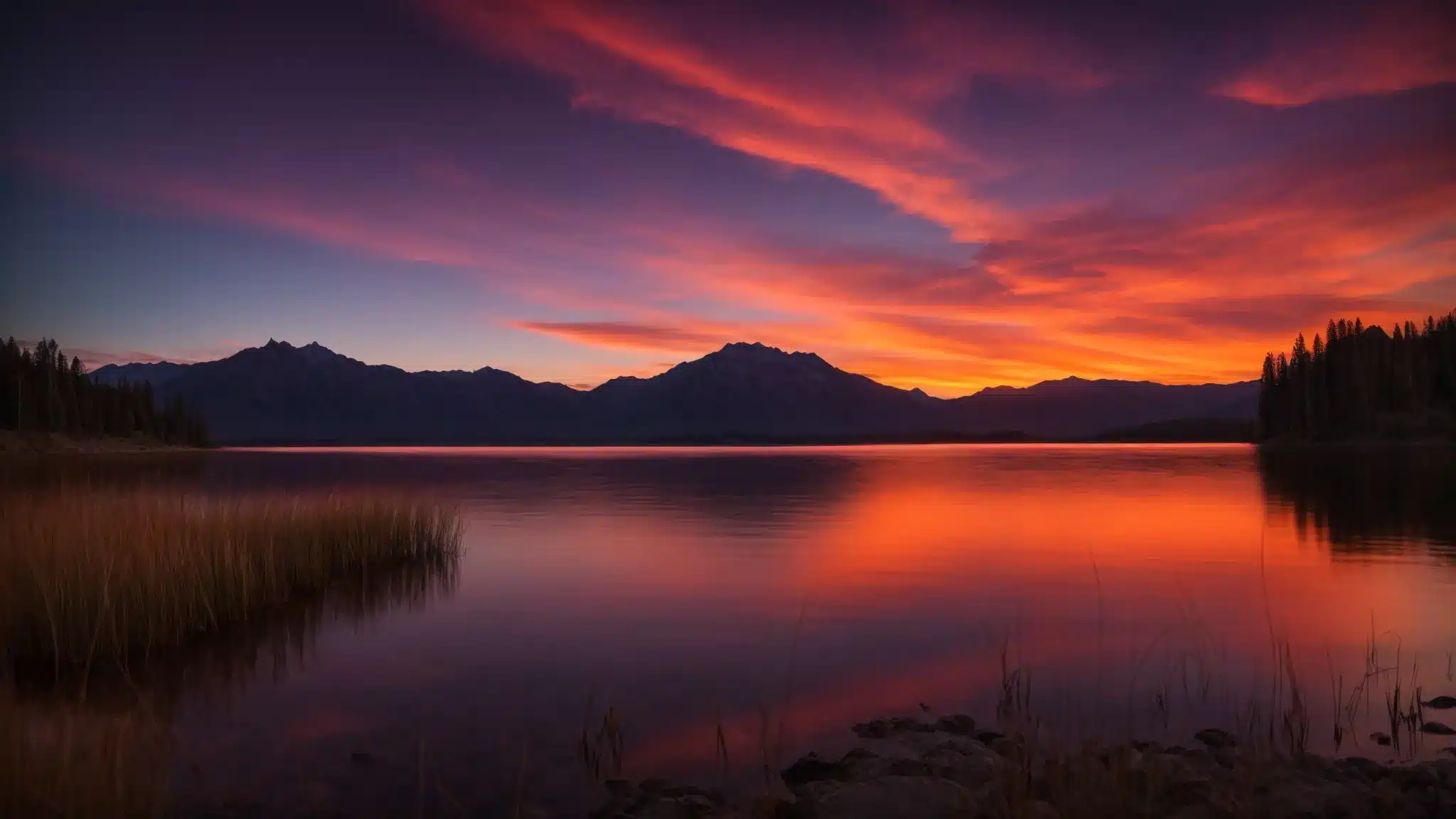 a breathtaking sunset casts vibrant hues over a tranquil lake, reflecting the fiery oranges and deep purples of the sky, while silhouetted mountains rise majestically in the background.